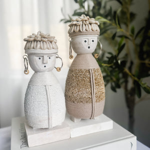 small white and medium beige dolls with macramé bodies, shell hats and beaded earrings on a wooden stand with an olive  tree in background