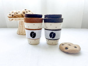 Two felt coffee cups with lids in mocha and latte colours with a chocolate chip felt cookies in rattan cake stand and one cookie beside coffee cup.