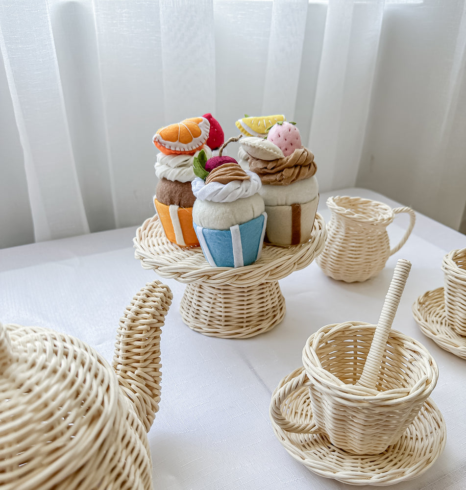 Set of three colourful felt cupcakes on a rattan tray surrounded by a milk jug, teapot, cups/saucers and spoons