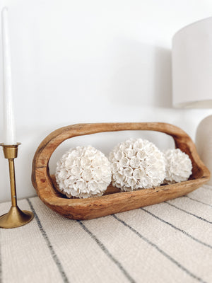 Wooden teak oblong basket with handle sitting on white shelf displaying three sizes of white shell balls with brass candlestick on one side and table lamp on other