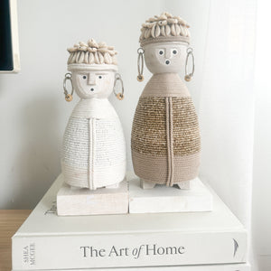 small white and medium beige dolls with macramé bodies, shell hats and beaded earrings on a wooden stand with sitting on a designer book