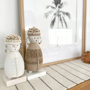 small white and medium beige dolls with macramé bodies, shell hats and beaded earrings on a wooden stand with a palm tree picture in background