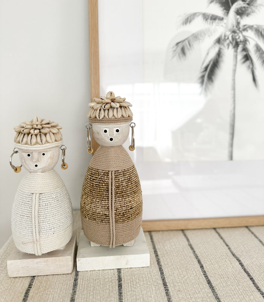 small white and medium beige dolls with macramé bodies, shell hats and beaded earrings on a wooden stand with a palm tree picture in background