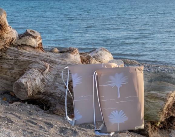 Beige tote bag with palm tree design on a beach with driftwood and ocean in the background