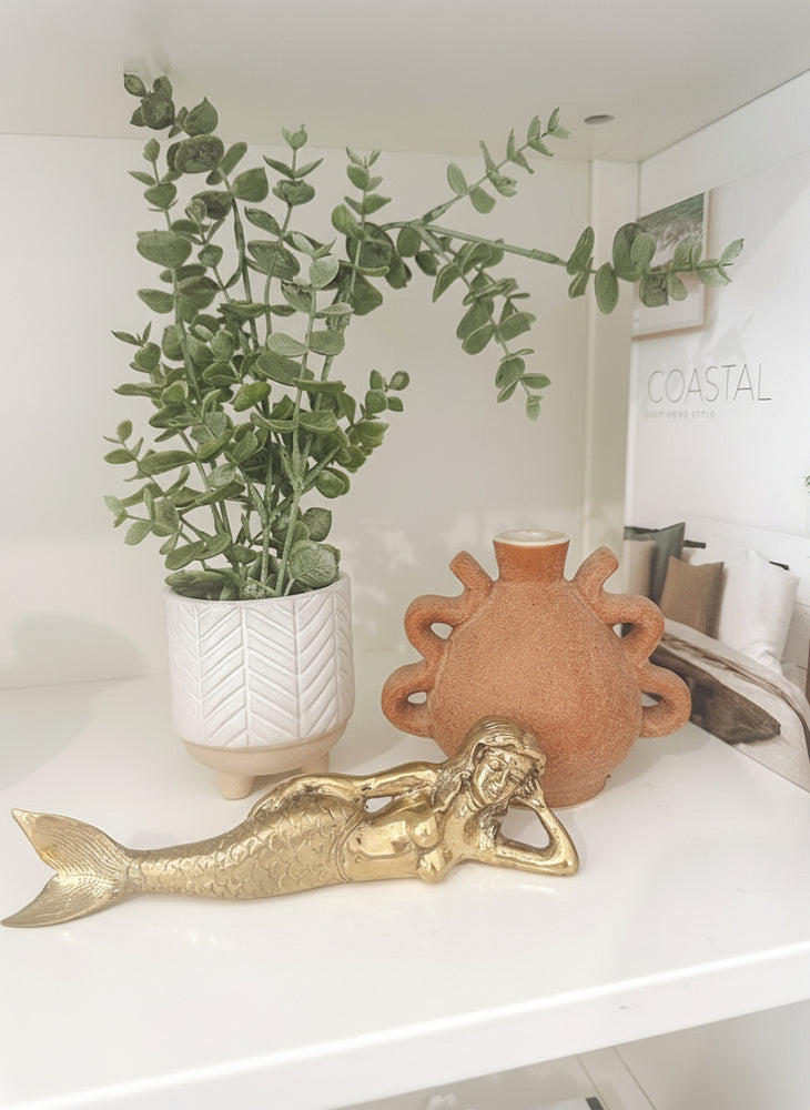Decorative items including a gold brass mermaid figurine, white pot with green plant, and terracotta sun-shaped vase on a shelf.