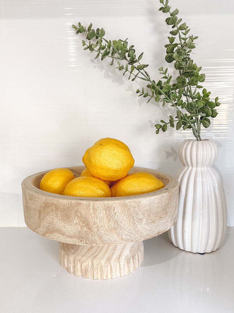 round paulownia wooden bowl on stand containing lemons displayed with white vase and olive plant