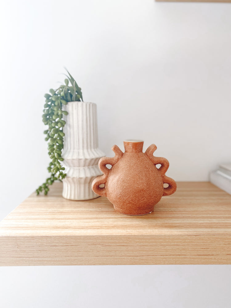 Small textured ceramic vase with two handles on each side displayed on a shelf with white vase